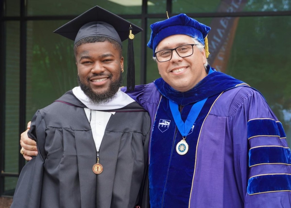 Luis Almeida standing with Donovan Bryan at graduation, both dressed in academic regalia and smiling for the photo.