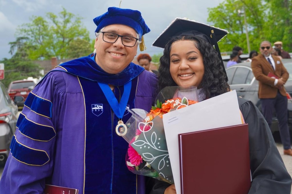 Luis Almeida standing with Ty Williams at graduation, both dressed in academic regalia and smiling for the photo.